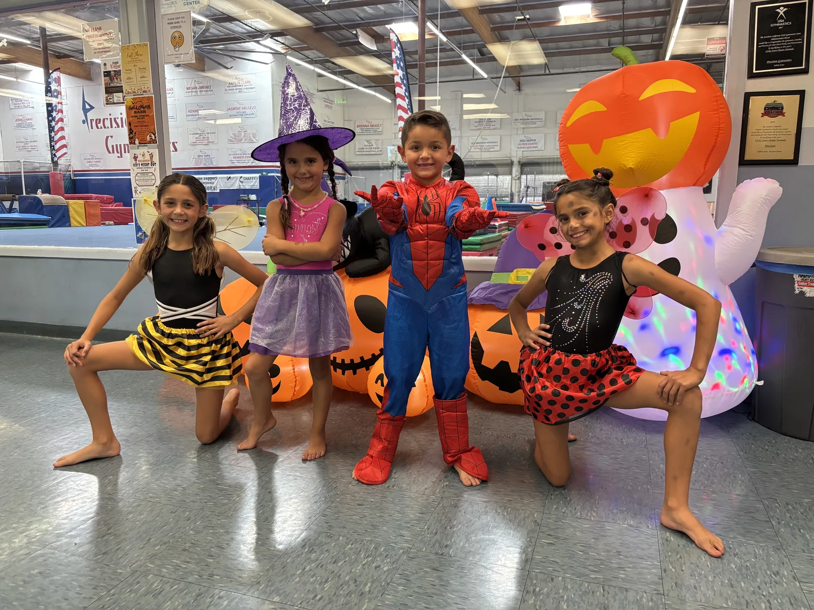 Four children in Halloween costumes — a bee, a witch, Spider-Man and a ladybug — posing in front of inflatable pumpkins at Precision